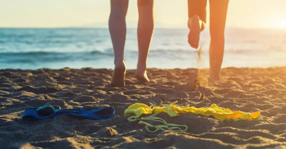 Two people walking barefoot on a beach at sunset, with beach clothes lying on the sand in the foreground and the sea in the background